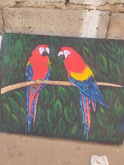 The image shows a painting of two macaws perched on a branch, set against a background of green leaves, likely representing a jungle or rainforest environment. The macaws are brightly colored, with red, yellow, and blue feathers. The painting appears to be resting against a rough, textured wall, possibly made of concrete or stone. The surface beneath the painting appears to be ground with a pale item. The painting could be in El Dátil, Venezuela.