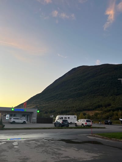 This image depicts an outdoor scene in Nordkjosbotn - Gárgán, Norway, at dusk. The sky above transitions from light blue at the zenith to pale yellow and pink hues near the western horizon, with scattered wispy clouds. A massive, densely forested mountain, predominantly dark green, dominates the background, rising sharply.
In the mid-ground on the left, a modern, single-story commercial building with a grey facade is visible. It features a prominent, illuminated neon green sign displaying "APOTEK 1" and a green cross emblem, indicating a pharmacy. A second green cross emblem is on the building's right side. A red and white flag flies from the building's roof, partially visible above the "APOTEK 1" sign. A white SUV, possibly a Subaru Outback, is parked directly in front of the pharmacy, facing left. A poster is visible within the building's front window to the left of the SUV.
The immediate foreground consists of a wet asphalt surface with multiple puddles reflecting ambient light. A concrete curb separates this area from a paved parking lot. In the parking lot, from left to right, are a large white recreational vehicle (RV) or motorhome, a dark grey SUV (possibly a Mazda CX-5), and a white pickup truck with a bed cap, potentially a Volkswagen Amarok.
Further to the right, beyond the white pickup truck, two individuals are present: one person is riding a bicycle, and another person is walking alongside them. Both appear to be dressed in dark clothing. A dark sedan or SUV is parked further down the road in the far right background. A tall street light pole is also visible on the far right edge of the image. A dark fence line runs horizontally across the mid-ground, separating the parking area from the base of the mountain. Green grass is visible on the right side of the paved area. Reflective road markers, some appearing blue and green, are present on the pavement in the lower foreground.