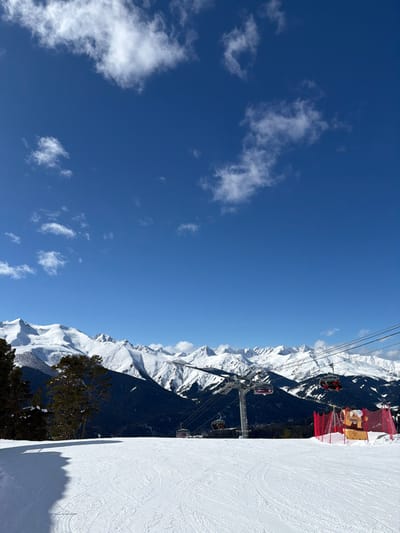 The image displays a winter mountainous landscape at a ski resort in Lunnaya Polyana, Russia. The upper two-thirds of the frame are dominated by a clear, bright blue sky with scattered white cumulus clouds. A range of snow-capped mountains stretches horizontally across the midground, featuring white snow cover and exposed rock. The lower slopes of these mountains are covered with dense, dark green coniferous forests.
In the foreground, a wide, groomed ski slope is visible, showing numerous ski tracks in the snow. A large, dark shadow from an unseen object or terrain feature extends across the left portion of the foreground snow. On the left side of the frame, dark green coniferous trees mark the edge of the ski area.
In the midground, a ski lift system is operational, featuring metallic support towers and several maroon-colored gondola cabins suspended by cables. A prominent ski lift tower stands in the center-right of the midground, with gondolas visible both to its left and right. To the far right foreground, a section of bright red safety netting is erected, behind which a small, light-colored structure with a cartoon-like illustration is positioned. No individuals are distinctly visible.