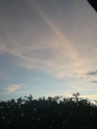 The image shows a sky with clouds and a contrail from an airplane. There is a tree with dark foliage visible on the right side of the image. In the upper left quadrant, there are two green lines, possibly from a laser pointer. The location is potentially in the city of Ub Municipality, Serbia, based on the image's source.