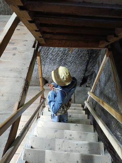 A person wearing a yellow hat and denim jeans sits on a set of wooden stairs inside the Turda Salt Mine in Romania. The person is facing away from the camera, looking down the stairs. The stairs are made of light-colored wood and appear to be old and worn. The walls of the mine are rough and textured, typical of a salt mine. Wooden railings are present on either side of the stairs. Some writing is visible on the wooden beams above the stairs.