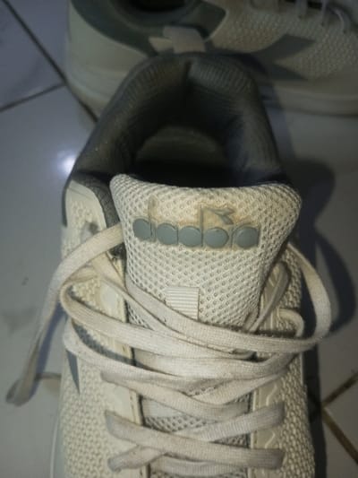 A close-up shot reveals a white Diadora sneaker with a mesh upper. The laces are untied and slightly soiled. The shoe's tongue features the brand's logo in gray. The inner lining of the sneaker appears gray and worn. Another similar sneaker, possibly the pair, is partially visible in the background, resting on a light-colored tiled surface. The overall impression is of a well-used and possibly dirty shoe.