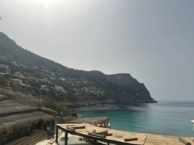 The image captures a scenic view in Port D'Andratx, Spain. A hillside densely populated with white-colored buildings is situated along the coast, transitioning into rocky terrain and cliffs that meet the sea. The turquoise water contrasts with the gray, overcast sky. In the foreground, a wooden structure with rolled bundles is present. A thatched-roof building is partially visible to the left. The scene appears to be a coastal construction or renovation area, with materials and ongoing work evident.