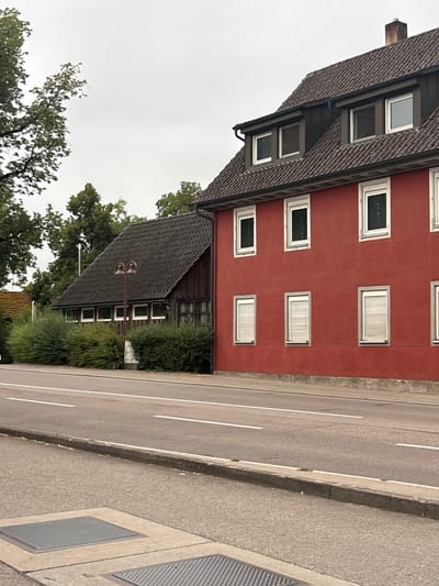 In Untermünkheim, Germany, a street scene unfolds. On the right, a red building with a dark tiled roof features multiple windows, some with closed shutters. Adjacent to it on the left is a lower, dark-tiled roof structure made of wood, also with several windows. Both buildings are bordered by green bushes. A paved road with white lane markings runs in front of the buildings, separated by a concrete curb. Two metal manhole covers are visible on the paved sidewalk area in the foreground. Trees are present in the background, and the sky is overcast.