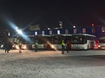 The image displays a nighttime scene in Yukhta, Russia, characterized by an outdoor, open area with a ground surface appearing to be covered in snow or frost. Multiple white and light-colored passenger buses are positioned across the midground, with several exhibiting illuminated headlights. In the background, a long, dark blue modular structure, resembling stacked shipping containers or prefabricated units, is visible, featuring numerous illuminated windows. A white van is partially observable on the far right side of the frame. Approximately six to eight individuals are present on the ground, with at least three distinctly wearing high-visibility yellow-green vests. Artificial light sources, including vehicle headlights and possibly overhead streetlights, generate significant lens flares in the upper-left and center portions of the image, illuminating the immediate foreground and vehicles. Distant industrial structures with scattered lights are faintly visible against the dark sky behind the modular building.