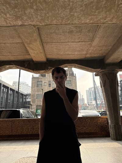 A medium shot depicts a person with short brown hair wearing a black top, standing in an architectural space. The person has one hand near their face, possibly holding something. The architectural space appears to be an outdoor corridor or alcove with a concrete ceiling and textured wall to the left.
Outside the corridor, there's a cityscape featuring buildings of various heights, some with multiple windows. The sky is overcast. A vehicle, possibly a car, is partially visible on the right side of the frame. The location is Hongkou District, China.