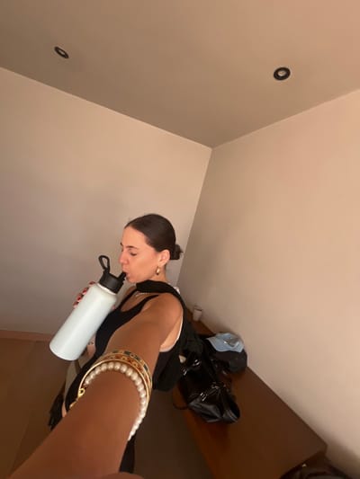 A woman is holding a white water bottle to her lips. She has dark hair in a bun, gold hoop earrings, and is wearing a black top. Her arm, adorned with layered bracelets, is extended towards the camera. In the background, a bed or ledge holds a black tote bag and other items. The room has light beige walls, and there are two circular dark objects on the wall near the ceiling.