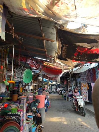 This is an outdoor market scene in the city of Gedang, Indonesia. The market is covered by a large, translucent canopy made of fabric stretched over a wooden frame. Various stalls are set up along a paved walkway. On the left side of the image, there are numerous household items displayed, including plastic containers, buckets, cleaning supplies like brooms, and a green dish rack. A woman wearing a blue headscarf and a mask is sitting among these items, facing a pile of goods. A calico cat is near her feet. Further down the walkway, another woman wearing a brown headscarf and a striped shirt is riding a white scooter. She is looking forward and appears to be moving through the market. Several other people and motorcycles are visible in the background, indicating a bustling environment. The stalls are filled with a variety of goods, including clothing on the right side of the image, with garments like patterned dresses and plaid shirts hanging on display. The overall impression is of a lively, somewhat cluttered marketplace.