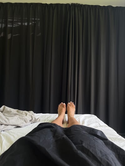 The image presents a first-person view from a supine position in a bed. In the immediate foreground, a black duvet covers the lower portion of the subject. Directly beyond the duvet, the lower legs and feet of an individual are visible, resting on white bed linens. The feet have white-painted toenails, and a light-colored anklet is visible on the right ankle. To the left of the legs, a crumpled light grey sheet or blanket is present on the white bed sheets. The entire background is occupied by dark, full-length curtains, likely black or very dark grey, which obscure any external view. Faint, lighter patterns are discernible through the upper left section of the curtains, indicating a window behind them. The specific location in Skanderborg, Denmark, cannot be determined from the visual information provided in the image.