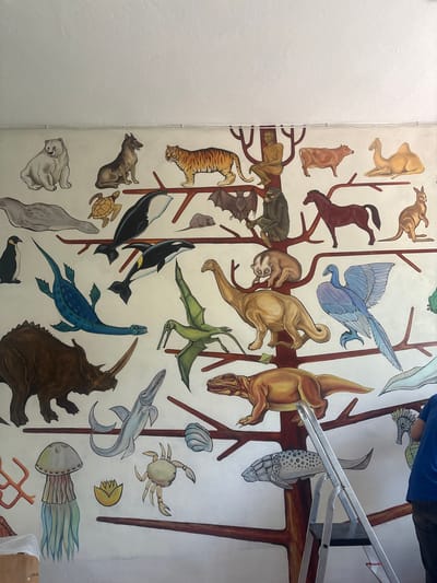 The image depicts a mural on a white wall, possibly in a public building or school in Bozhurishte, Bulgaria. The mural is a stylized tree of life, featuring various animals painted on its branches. Animals depicted include a polar bear, a wolf, a tiger, a seal, a penguin, a turtle, an orca, a plesiosaur, a triceratops, a bat, a chimpanzee, a horse, a kangaroo, a slow loris, a theropod dinosaur, a bird (possibly Archaeopteryx), a bird-like dinosaur, a pterosaur, a cow, a camel, a jellyfish, a crab, a fossilized ammonite, and a seahorse. A silver stepladder stands in the foreground, with a person in a blue shirt and dark pants visible on the right side of the frame, appearing to be working on or observing the mural. The environment is indoor, with a white ceiling visible at the top of the image.