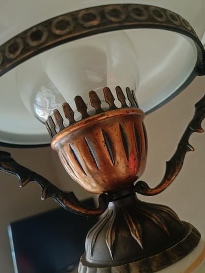 This is a close-up, low-angle shot of the decorative metalwork and glass shade of an antique lamp. The lamp features a prominent copper-colored metal cup with a fluted design and decorative accents, holding a clear glass lightbulb. This cup is supported by ornate, dark metal arms that curve outwards. Above the cup, a wide, white lampshade with a decorative metal rim is visible. The background is softly blurred, showing a hint of a dark object, possibly a television, and a neutral-colored wall. The lighting is soft and natural, highlighting the textures and finishes of the lamp. The specific location in Niasvizh, Belarus is not discernible from the image.