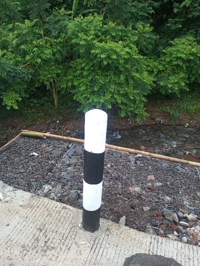 A black and white striped pole stands on a concrete surface near a stream. The stream is lined with rocks held in place by wire mesh and bamboo poles. Green trees and bushes densely cover the background. A small amount of litter is visible along the stream bank.