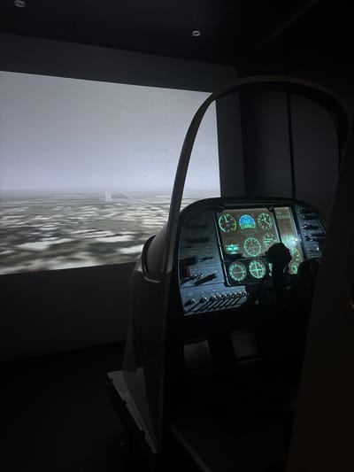 This image depicts the interior of a flight simulator located in Port Alfred, South Africa. The scene is observed from a vantage point within the cockpit, which occupies the right side and foreground of the frame. The simulator's instrument panel is brightly illuminated with glowing green and blue digital and analog gauges, including multiple circular dials, an array of toggle switches, and a partially visible control yoke. The surrounding room is dark. A large projection screen on the left and in the background displays a simulated exterior environment: a flat, textured landscape under a uniformly grey, overcast sky, with a clear runway visible in the distance. Small white speckles are present across the projected sky, indicative of simulated precipitation or screen artifacts. No individuals are visible operating or observing the simulator.