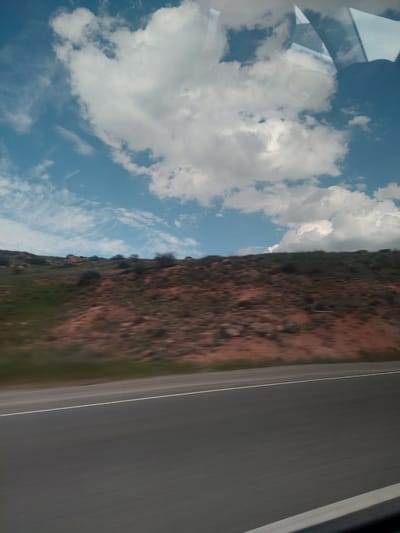 The image captures a landscape scene, likely viewed from a moving vehicle, as suggested by the motion blur on the road and reflections on a window pane. The photograph appears to have been taken with the camera rotated, presenting the road and land vertically, and the sky horizontally.

When re-oriented, the scene depicts a wide, grey asphalt road across the bottom of the frame, featuring a white solid line along its edge. The blurred appearance of the road indicates movement. Adjacent to the road, a rugged, reddish-brown hillside or mountainous terrain dominates the mid-ground, partially covered with patches of green vegetation and scattered shrubs. Above this natural landscape, a bright blue sky is filled with a dynamic array of white clouds, ranging from large, fluffy cumulus formations to more delicate, wispy streaks.

In the upper left corner, reflections are visible on what appears to be a vehicle window, showing indistinct shapes possibly from the car's interior or blurred external elements. The clear blue sky and strong illumination suggest it is daytime, under fair weather conditions. The composition evokes a sense of travel through an open, natural environment. No discernible text is visible within the image. The provided location context is Tigranashen, Armenia.