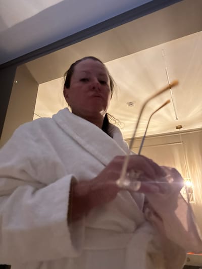 A low-angle shot features a woman wearing a white robe, holding glasses. Her face, partially obscured by the glasses, is visible. The background includes a white ceiling with recessed lighting. A dark-colored wall is visible on the left edge of the frame. The view is looking upwards at the woman.