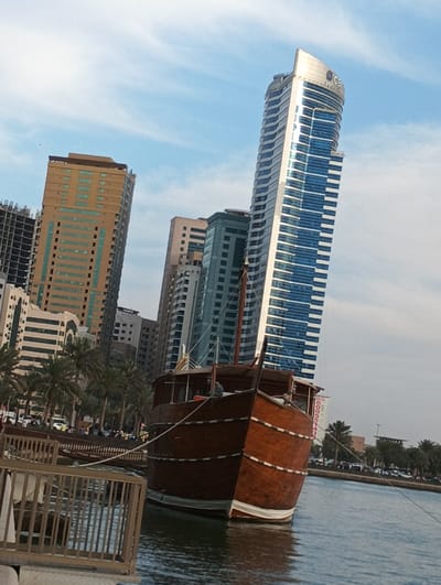 A wooden dhow boat is docked in the water, in front of a large, modern skyscraper. The skyscraper, with reflective glass panels, is located in Sharjah, United Arab Emirates, at the coordinates 25.334, 55.388. Behind the boat, a variety of other buildings, including several high-rises, can be seen. The boat is secured to a dock. A small crowd of people can be seen walking along the shore. The sky is bright blue with a few wispy clouds. A palm tree grows behind the boat. A second palm tree, along with a row of smaller trees, grow on the shore. Several people are standing at the edge of the water. The water is calm. The scene is a mix of modern and traditional architecture.