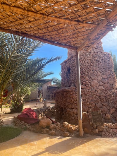 A low-angle shot reveals a sunny outdoor scene. A woven wooden structure partially frames the view, contrasting with a rock wall made of rounded stones. Palm fronds sway in the center, with a glimpse of a building in the background. A red cloth drapes over stones near a tiled surface and small patch of grass. The clear blue sky is visible through the gaps.