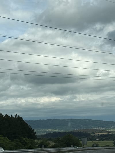 The image captures a cloudy sky over a landscape that appears to be Staro Selo, Bulgaria. In the foreground, a highway barrier runs horizontally across the scene. A white car is partially visible on the left side of the highway. Beyond the barrier, the terrain slopes downwards, revealing a dense forest of dark green trees on the left. In the middle ground and background, rolling hills are covered in lighter green vegetation and scattered trees. Distant buildings are visible as small clusters within the landscape. Power lines are stretched across the upper portion of the image. The overall atmosphere is overcast, with diffused light characteristic of a cloudy day.