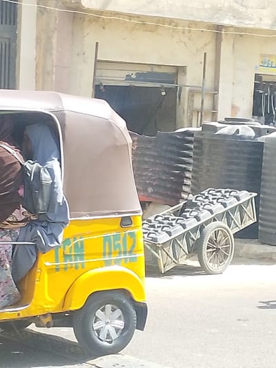 An outdoor street scene in Darmanawa, Nigeria, depicts a yellow three-wheeled auto-rickshaw in the left foreground and a two-wheeled utility cart to its right. The auto-rickshaw features a brown canvas roof and displays "TNN 050Z" in green lettering on its side panel above the rear wheel. At least two occupants are visible within the auto-rickshaw; one wears a brown garment, while another wears a light blue hijab and grey attire, carrying a grey bag. The utility cart, constructed with dark-colored framing, is laden with approximately 15-20 black plastic containers, each fitted with a top handle, consistent with water jerrycans. In the background, a light-colored, weathered building with a dark rectangular entrance is present. Multiple large, black cylindrical plastic water storage tanks are stacked near the building, to the right of the cart. A thin white line, possibly a utility cable, spans horizontally across the upper portion of the background. The ground consists of a light-colored, possibly unpaved, street surface.
