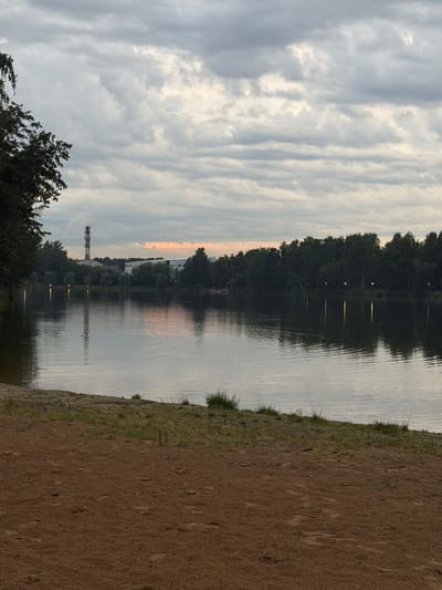 The image depicts a tranquil scene at the Malakhovka water reservoir in Malakhovka, Russia. The foreground is a sandy beach with scattered footprints and sparse green vegetation. Beyond the beach, a calm body of water reflects the overcast sky, which is filled with a mixture of grey and white clouds, with a hint of soft orange light near the horizon. The far bank of the reservoir is lined with a dense row of trees. In the distance, several lights are visible along the tree line, casting reflections on the water. To the left of the frame, a tall, red and white striped tower, possibly a communication tower, stands against the sky, accompanied by some low-rise buildings. The overall atmosphere is serene and subdued, suggesting either early morning or late evening. There are no discernible people or animals in the frame.