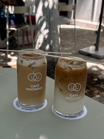 The image captures two iced beverages, likely coffees or lattes, placed on a light-colored outdoor table. Each drink is served in a clear glass bottle with a white paper straw inserted. The bottles are prominently branded with a unique logo featuring two intertwined face icons, one smiling and one frowning, alongside the text "CAFÉ SINONIMO".

The upper drink displays a distinct two-toned layer, with darker coffee settling below a creamy white top layer, suggesting an iced latte. The lower beverage, however, appears more uniformly blended to a light brown hue. Both bottles contain visible ice cubes.

Underneath each bottle is a round white coaster, also printed with the name "Cafe Sinonimo" repeated along its edge. The table surface is a pale, possibly light green or off-white color. The background is softly blurred but reveals an outdoor environment, with dappled sunlight and shadows indicating the presence of trees or overhead structures, consistent with a cafe patio setting.

The bright, natural illumination suggests it is daytime, likely morning or early afternoon, suitable for enjoying cold beverages outdoors in Guadalajara, Mexico. The scene evokes a sense of relaxation and casual enjoyment at a coffee establishment. No individuals are visible, but the presence of two drinks implies a shared experience or an individual indulging in multiple refreshments.