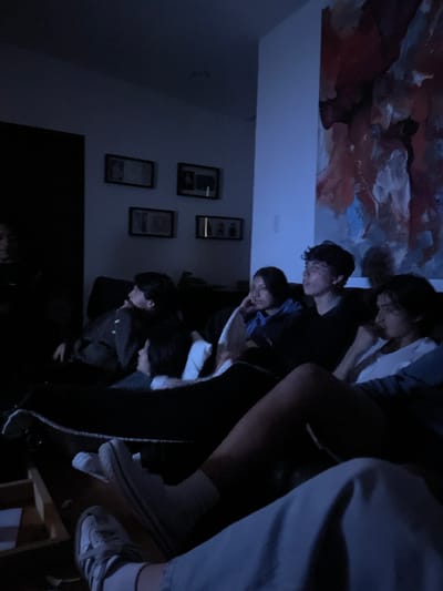 The image appears to be taken indoors in a dimly lit room, perhaps at night. A group of approximately six young individuals are clustered together, partially obscuring each other. They seem to be looking down, possibly at a shared screen or object out of view.
On the wall, there are framed pictures. Above the group, a colorful abstract painting is visible on the ceiling or upper wall. The location, El Oasis Valsequillo, Mexico, cannot be confirmed from visual details alone. The interaction appears casual, suggestive of a social gathering.