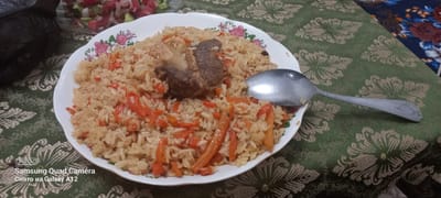 A close-up, overhead shot shows a plate of plov, a rice pilaf dish, served on a green tablecloth with an ornate floral pattern. The plov is a generous portion of rice mixed with carrots and small pieces of meat, with a large chunk of meat prominently placed on top. A metal spoon rests on the right side of the plate, angled towards the bottom right corner. In the upper left corner, a blurry glass bowl contains a salad of chopped tomatoes and onions, suggesting a meal is in progress. The scene appears to be indoors, likely a dining setting. The image also contains a visible watermark in the bottom left corner: "Samsung Quad Camera Снято на Galaxy A12," indicating the photo was taken with a Samsung Galaxy A12 phone. The lighting is artificial and somewhat dim, casting soft shadows. The overall impression is one of a home-cooked meal, possibly lunch or dinner, in Levakant, Tajikistan.