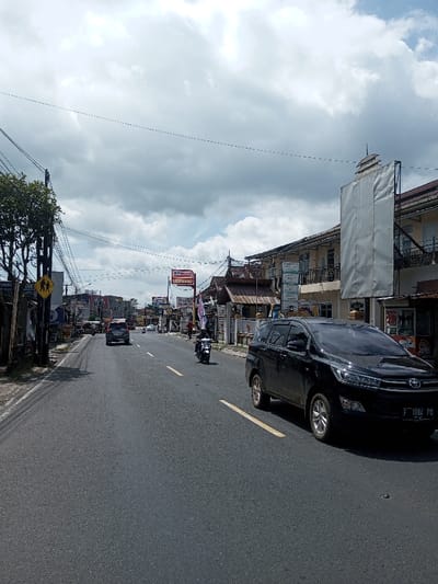 This image depicts an urban street scene. While the requested location is Gubbisamamidi, India, visual evidence such as the 'Alfamart' sign and the vehicle license plate 'F 1884 PQ' indicates the scene is located in Indonesia.
The scene shows a two-lane asphalt road, marked with dashed yellow center lines and a solid white line on the right shoulder. In the foreground, a dark-colored Toyota Innova (or similar SUV/MPV) is positioned on the right side of the road, displaying an Indonesian license plate "F 1884 PQ". Further down the road, a motorcyclist wearing a helmet is traveling towards the viewer in the left lane, while a dark-colored sedan or small SUV drives away in the same lane. Other vehicles are visible in the distant background.
To the left, a yellow diamond-shaped "school crossing" sign depicting two walking figures is affixed to a utility pole, alongside a leafy green tree. The right side of the street features a row of multi-story commercial and residential buildings. One building prominently displays an "Alfamart" sign, while another adjacent structure has a large white covering on its upper facade. The sky above is partly cloudy with significant grey cloud cover and patches of blue sky. Numerous overhead utility lines crisscross the street.