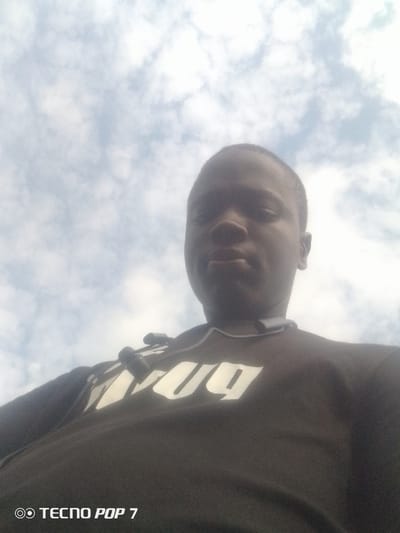 A low-angle, close-up selfie shows a young Black male from the chest up against a cloudy sky. The subject's head is tilted back, and his gaze is directed upwards. He is wearing a black t-shirt with large, stylized white lettering across the chest, partially obscured by the camera angle. A dark grey pair of earbuds with black tips rests around his neck. The lower portion of the image shows the text "TECNO POP 7" in white font. The image appears to have been taken outdoors in Eleweran, Nigeria, given the watermark.