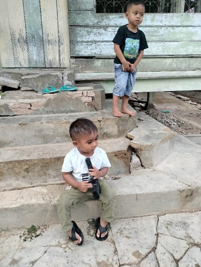 This image, captured in Cutneuheun, Indonesia, depicts two young males on and around concrete steps outside a structure with weathered wooden siding. In the foreground, a younger male child is seated on the lowest concrete step, wearing a white t-shirt, olive green pants, and black flip-flops. He holds a black, rifle-like toy or replica firearm in both hands, resting it vertically on his lap, and is looking towards the right side of the frame. A pair of black flip-flops is visible near his feet. In the background, an older male child stands barefoot on an upper concrete step, wearing a black t-shirt with a graphic print and light blue knee-length shorts. He faces right, looking away from the camera, with his hands clasped or adjusting his shorts at the waist level. A pair of teal flip-flops rests on a concrete step to the left of the standing child. The building behind them features vertical wooden planks with faded and peeling light green and white paint. The concrete steps exhibit cracks, damage, and exposed brickwork in various areas. The ground surface consists of cracked concrete and loose soil with sparse vegetation.