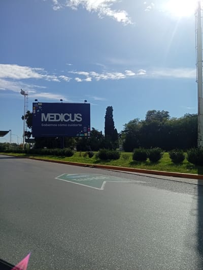 The image shows a Medicus billboard located at the Aeropuerto Internacional Ezeiza in Argentina. The billboard displays the Medicus logo and the slogan "Sabemos cómo cuidarte" (We know how to take care of you). The billboard is set against a backdrop of trees and a blue sky with scattered clouds. In the foreground, there is an asphalt road with a painted arrow pointing to the right. A light pole is visible on the left, and a taller metal structure is on the right side of the image. The environment is outdoors during daylight.