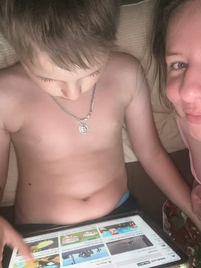 This image shows two people, likely a mother and child, lying down and looking at a tablet. The woman in the foreground is partially visible, with her eye and part of her face in focus. Her hair is blonde, and she appears to be wearing a pink garment. The child is positioned below her, with their torso and head visible. The child is also looking at the tablet, which is displaying the Roblox game platform. The screen shows various game titles in Russian, including "ТАНКИ!" (Tanks!), "Мир.io" (Mir.io), and others. The scene appears to be indoors, with the subjects lying on a couch or bed. The lighting suggests it could be daytime. The overall mood seems relaxed and casual, with the mother and child engaged in a shared activity. The time stamp on the tablet's screen indicates "20:56 06.27 черв," which translates to 8:56 PM on June 27th.