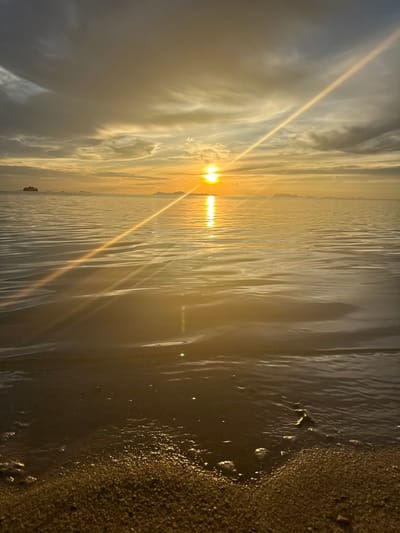 The image depicts a serene ocean sunset scene at Baan Pang Ka, Thailand. The sun, a bright orb of golden light, is low on the horizon, casting a shimmering reflection across the calm water. Wispy clouds stretch across the sky, tinged with hues of orange and grey, creating a dramatic backdrop. In the distance, a faint outline of islands or landmasses can be seen. The foreground is a sandy beach, with gentle waves lapping at the shore, creating a subtle foamy texture. The overall atmosphere is one of tranquility and natural beauty.
