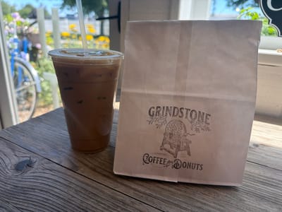 A close-up shot shows an iced coffee in a plastic cup with a straw and a brown paper bag with the "GRINDSTONE COFFEE AND DONUTS" logo on a weathered wooden surface. The background is blurred, featuring a bicycle, flowers, and a window with sunlight. The scene is outdoors, likely in a cafe or bakery setting within the Village of East Hampton, United States of America.
