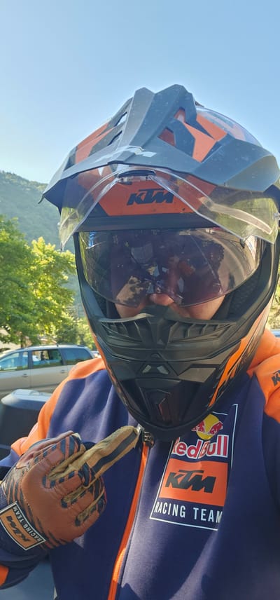 A person is wearing a full-face dirt bike helmet with an orange and black design and a clear visor. The helmet has a KTM logo on the front. The person is also wearing an orange and blue racing jacket with a Red Bull and KTM Racing Team logo. They are also wearing brown and black dirt bike gloves. The person is pointing with their right index finger. The background shows trees, a car, and a hilly landscape under a clear blue sky. The image appears to be taken outdoors on a sunny day, possibly in a rural or mountainous area. The location is Stara Kresna, Bulgaria.