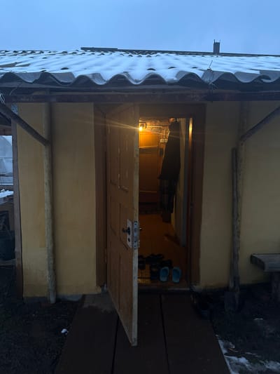 The image shows a small, one-story structure in Zapadnaya, Russia. The building has a corrugated roof covered in patches of snow. The walls are painted a faded yellow. The open doorway reveals a cluttered interior with a visible light source. Outside the doorway, there are some shoes and gardening tools. The ground is partly covered with dark soil and remnants of snow. The sky is a light blue.