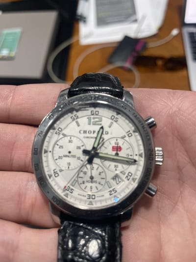 An overhead, close-up shot captures a person's left hand holding a Chopard chronograph watch indoors. The watch features a white dial with three subdials, luminous hour and minute hands, and a black crocodile-patterned leather strap. The silver-toned bezel is inscribed with a "TACHYMETER" scale and numbers such as 240, 200, 180, 100, 85, 80, and 75. Visible text on the watch face includes "CHOPARD," "CHRONOMETER," "20 MINUTES," and "SWISS MADE." A red square symbol is present on one of the subdials, and the number "12" is prominently displayed on the main dial. The background is softly blurred, showing hints of papers, cables, and electronic devices on what appears to be a desk or table surface. The image is taken under artificial indoor lighting, with no specific weather or time of day discernible. The setting is likely a home or office in Cuautitlán Izcalli, Mexico.