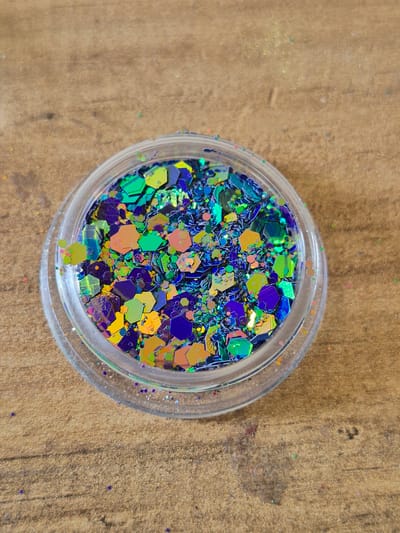 A clear, round plastic container filled with iridescent glitter is pictured, placed on a textured wooden surface. The glitter is a mix of hexagonal and irregular shaped pieces in vibrant shades of blue, green, purple, and orange, reflecting light with a metallic sheen. Some loose glitter particles are scattered on the wooden surface surrounding the container. The wooden surface has a visible grain pattern and appears to be a tabletop or a similar flat surface. The image is taken from a high-angle perspective, looking down onto the glitter container. The location is not specified within the image or its metadata.