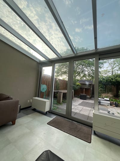 The image depicts the interior of a conservatory or sunroom. Light enters through the angled glass roof, reflecting the sky and clouds. The floor is tiled in a light color. Outside the large glass doors is a garden area with plants and a fence. A coiled blue garden hose is mounted on the wall inside the conservatory. Two white rectangular objects, possibly air conditioners or heaters, are visible. A brown rug lies near the entrance. A portion of furniture is visible, suggesting the space is used for seating. Based on the request, it is inferred that the location of the image is Oud-Beijerland, Netherlands.