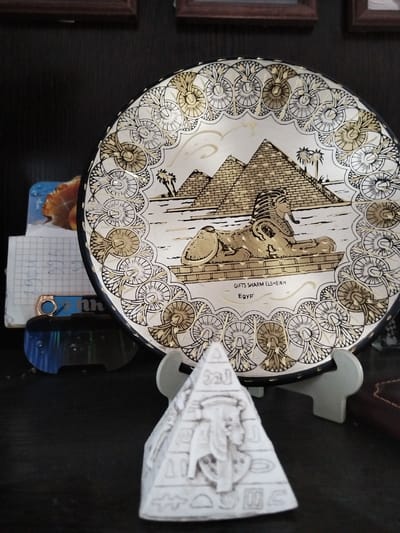 The image shows a decorative plate with an Egyptian theme, featuring pyramids and the Sphinx. The plate is white with black and gold accents and has a decorative border. It is propped up on a stand. In front of the plate is a small pyramid-shaped object with hieroglyphs. To the left of the plate are some papers, one with a grid pattern. The background appears to be a dark surface, possibly a shelf, with framed pictures visible at the top. The scene is dimly lit. There are no people or any indication of the city of Bezlesnyy, Russia.