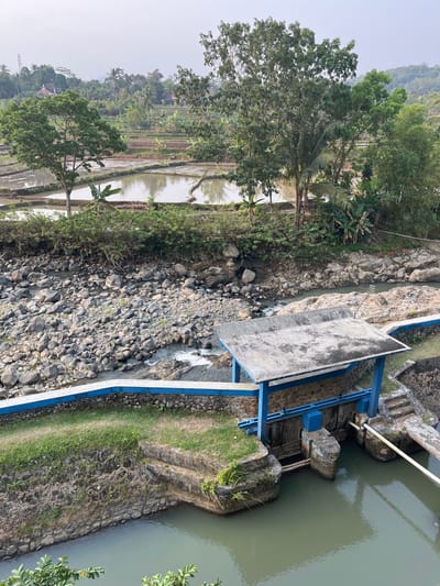 An aerial shot captures a serene landscape in Krasak, Indonesia, featuring a waterway with a small dam structure. The dam, built with concrete and supported by blue metal beams, has a weathered concrete roof. Water flows through a channel adjacent to the dam, which appears to be a part of a water management system. The surrounding environment is lush and green, with terraced rice paddies visible in the background, indicating agricultural activity. Trees of various sizes and types dot the landscape, adding to the natural beauty of the scene. The sky is overcast, creating a soft, diffused light. The waterway itself is lined with rocks, and the water is a muted green color.