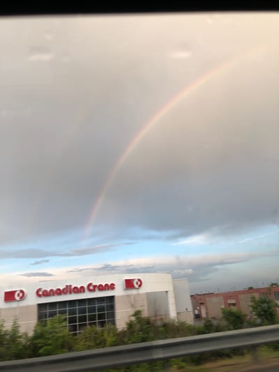 The image captures a partial view of a rainbow arcing across a cloudy sky. Below the sky, a white building with red signage that reads "Canadian Crane" is visible. The building has a large glass-paned section. To the right of the "Canadian Crane" building, another brick building with boarded-up windows can be seen. The scene appears to be along a roadway, with a guardrail in the foreground. The location is identified as Barrie, Canada.