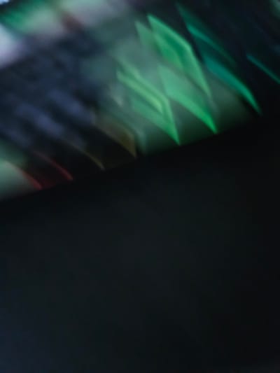 The image is an abstract, blurry close-up of light patterns. Streaks and shapes of green and dark blue dominate the upper portion of the frame, suggesting refracted or reflected light. There are also faint hints of red and yellow. The lower half of the image is a solid, dark, out-of-focus area. There are no identifiable people, objects, or environmental features that would suggest a specific location, including Versailles, France.