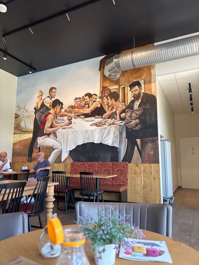 The image depicts the interior of a restaurant or cafe in Elsenfeld, Germany. A large mural covers the wall, portraying a scene of people gathered around a table, reminiscent of "The Last Supper." The mural features figures in historical attire, with a central focus on a communal meal. In the foreground, patrons are seated at tables, engaged in dining or conversation. The restaurant's decor includes wooden furniture, upholstered seating, and decorative elements like a small potted plant. A large, corrugated ventilation duct runs along the ceiling, partially obscuring the mural in some areas. The lighting appears to be a combination of track lighting and ambient room light, creating a well-lit environment.