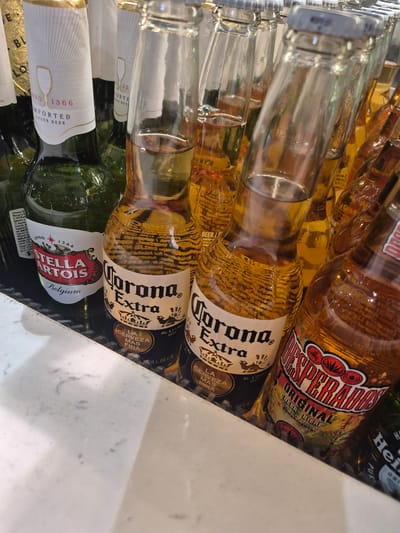 The image shows a close-up, eye-level view of several bottles of beer, primarily Corona Extra and Desperados, displayed in a refrigerated unit. The bottles are arranged closely together, with some partially obscuring others. The lighting appears to be artificial and overhead, casting reflections on the glass bottles. The background is dark, suggesting the inside of a cooler or refrigerator. The foreground consists of a white, slightly textured surface, possibly the edge of the display unit or a counter. The labels on the bottles are clearly visible, indicating brand names and some descriptive text. One bottle of Stella Artois is also visible. There are no people or animals in the frame, and no discernible activities are taking place. The overall impression is of a retail display of alcoholic beverages, likely in a bar, restaurant, or shop. The location is identified as Luqa, Malta.