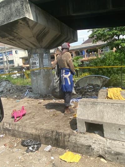 The image depicts an outdoor urban scene under a concrete bridge or overpass structure in Jakande, Nigeria, on a sunny day.
In the foreground, a dark-colored curb separates the immediate viewer's space from a dirt and gravel ground heavily littered with various debris including plastic bottles, wrappers, and a crumpled yellow item. To the left, a bright pink child's toy or bag with a handle lies on the ground. On the right, large, weathered concrete barriers are present, one with a rectangular cutout.
In the mid-ground, a single individual is seen from behind, standing on the dirt ground. The person wears a light brown, torn, open-knit long-sleeved top, dark trousers, and dark slip-on sandals. A blue garment with a large white "R" is tied around their waist, and a red and white striped fabric covers their head. The individual is carrying a large, dark grey/black plastic bag over their right shoulder. To the right of the individual, a large, inverted, broken dish-like object (possibly a satellite dish) filled with refuse is visible, with a yellow fabric item draped over an adjacent concrete barrier.
A weathered concrete support pillar for the overhead structure stands centrally, covered in posters, including one mentioning "PCI CONTRACTORS," "JAMB SSCE," and a partial phone number "3464061." A yellow tape or rope runs horizontally across the mid-ground, behind the individual and the concrete pillar.
In the background, green trees and foliage are visible, partially obscuring multi-story buildings. One building features a red roof, multiple balconies, and an orange facade. Another structure with a blue banner is visible further left. The overhead concrete bridge structure shows signs of weathering and staining.