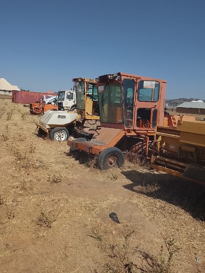 The image depicts an outdoor storage or staging area for heavy machinery in Bassa, Nigeria, under a clear, cloudless blue sky. The ground is composed of dry, reddish-brown earth with sparse, desiccated yellowish-brown vegetation.
In the midground, several pieces of heavy equipment are parked. The most prominent machine in the right foreground is an orange-red road paver or similar construction vehicle, featuring an enclosed cabin with multiple glass windows; a "FOR SALE" sign is visible on its front window. Its wheels appear dark and somewhat recessed. To its left and slightly behind it is a yellow piece of equipment, possibly a road sprayer or maintenance vehicle, with a white, tank-like component at its front labeled "ROSCO" in black lettering, and equipped with white-rimmed wheels. Behind these, a white truck cab is partially visible, along with another yellow machine and an orange piece of equipment that shows a partial boom or arm structure.
In the background, to the left, a long row of red shipping containers is visible, stacked horizontally. Beyond them, a white-domed building with a red roof structure is partially obscured. To the right, a distant rocky, hilly outcrop dominates the horizon, beneath which several light-colored, simple buildings with dark roofs are situated. No individuals are present in the scene. Shadows from the machinery extend across the dry ground towards the lower right.