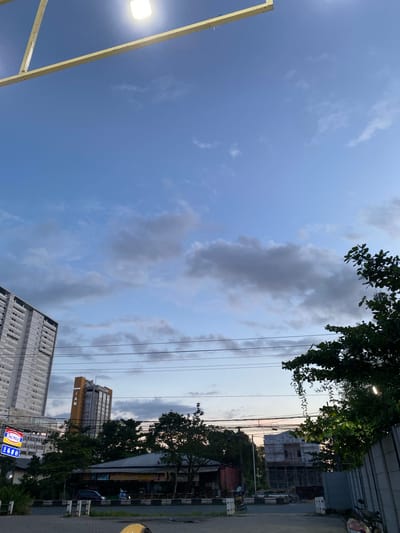 The image captures a dusk scene in Sungai Lakum, Indonesia, characterized by a vast expanse of cloudy blue sky above a low-rise urban landscape. In the foreground, a yellow metal structure with a bright light fixture is visible against the sky. Below, the cityscape unfolds with a tall, modern apartment building on the left, its windows and balconies distinct. Adjacent to it, a shorter, yellow-accented building with "GRAND IAN" signage stands out. A dense line of trees partially obscures a row of low-lying buildings and structures, some of which appear to be shops or businesses. Electrical wires crisscross the mid-ground. A street with parked cars and a few pedestrians is visible in the lower portion of the image, suggesting a public thoroughfare. Construction scaffolding is present on a building in the background on the right. The overall impression is one of an active urban environment transitioning into evening.