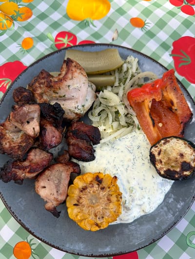 A plate of food is centered on a table covered with a green and white checkered tablecloth with tomato and orange graphics. The plate, which is gray and speckled, holds a variety of cooked items: grilled red pepper, grilled zucchini, grilled corn, pickles, an onion salad, a white sauce, and grilled meat. The meat pieces vary in color from light to dark brown. The onion salad appears to be dill-based. The background setting or location is not explicitly discernible. There is no information to suggest this is in Plieņciems, Latvia.