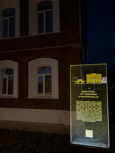 The image displays a dark brick building, identified as a historic house, illuminated by a glowing informational sign at night in Nikolo-Beryozovka, Russia. The building features a dark red brick facade with several windows, each framed by white, ornate arch-shaped sills and surrounds. The sky above is dark blue, indicating nighttime. Below the building's foundation, there is a strip of light-colored ground, followed by dark, sparse vegetation, likely grass.
The prominent feature is a tall, transparent, rectangular sign, glowing with a yellow light, positioned in front of the building. The sign's text, clearly visible, reads "ДОМ КУПЦА С.Н. ДЕДЮХИНА" (Merchant S.N. Dedyukhina's House) and "ул. Большая Купеческая, д.84" (Bolshaya Kupecheskaya Street, house 84). It includes two stylized illustrations of the building, with the number "84" placed next to one.
The detailed text on the sign describes the estate of Stepan Nikiforovich Dedyukhin, noting his arrival in Nikolo-Beryozovka around 1879. It highlights his business ventures in bread and timber trade, owning steamboats and barges. Dedyukhin was also a church elder, hosted Grand Duchess Elizabeth Fyodorovna, bishops, and governors, and oversaw significant renovations and new construction at the church between 1902 and 1904. After his departure in 1918, the house hosted N.K. Krupskaya in 1919 and subsequently served as a pedagogical school (from 1919), the Nikolo-Beryozovsky orphanage (from 1956), and a district hospital (from 1972). A QR code is also visible on the sign. The scene captures a historical landmark, providing rich contextual information about its past.